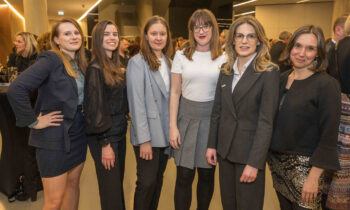 Sechs Frauen in Geschäftskleidung stehen zusammen und lächeln bei einer Indoor-Veranstaltung, bei der sich die Leute im Hintergrund mischen.