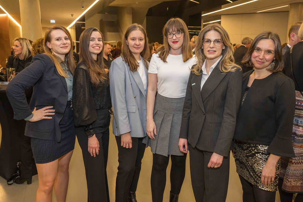 Sechs Frauen in Geschäftskleidung stehen zusammen und lächeln bei einer Indoor-Veranstaltung, bei der sich die Leute im Hintergrund mischen.