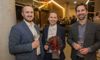 Drei Personen in formeller Kleidung lächeln bei einer Veranstaltung und halten Gläser mit Wein; die Frau in der Mitte hält eine Trophäe mit der Aufschrift "Spirit of Women Award".