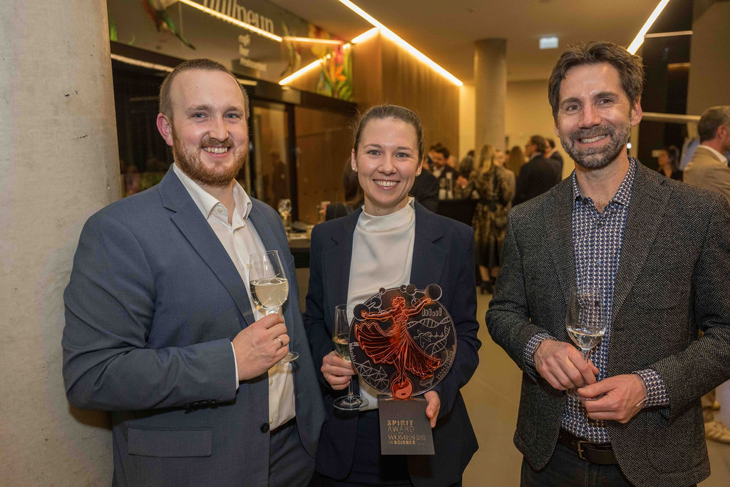 Drei Personen in formeller Kleidung lächeln bei einer Veranstaltung und halten Gläser mit Wein; die Frau in der Mitte hält eine Trophäe mit der Aufschrift "Spirit of Women Award".