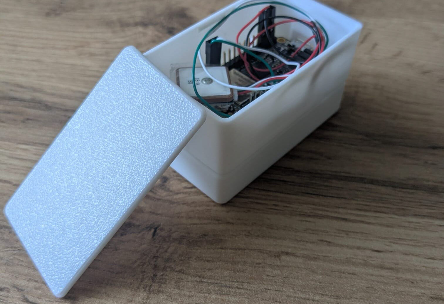 Ein kleines weißes Plastikkästchen mit abgenommenem Deckel, in dem elektronische Bauteile und Drähte zu sehen sind, steht auf einer hölzernen Unterlage.