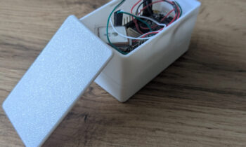 Ein kleines weißes Plastikkästchen mit abgenommenem Deckel, in dem elektronische Bauteile und Drähte zu sehen sind, steht auf einer hölzernen Unterlage.