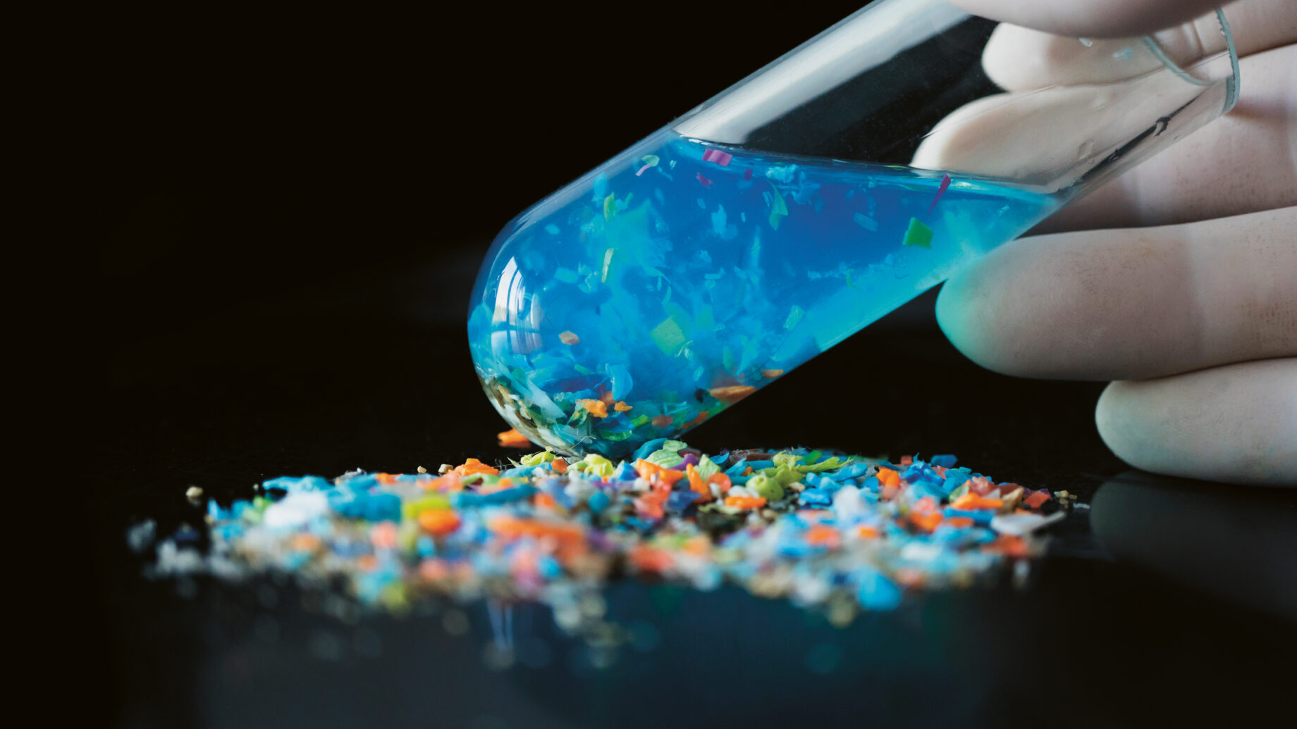 Eine behandschuhte Hand kippt ein Reagenzglas mit blauer Flüssigkeit und buntem Mikroplastik, das sich auf einer schwarzen Fläche ergießt.