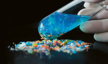 Eine behandschuhte Hand kippt ein Reagenzglas mit blauer Flüssigkeit und buntem Mikroplastik, das sich auf einer schwarzen Fläche ergießt.