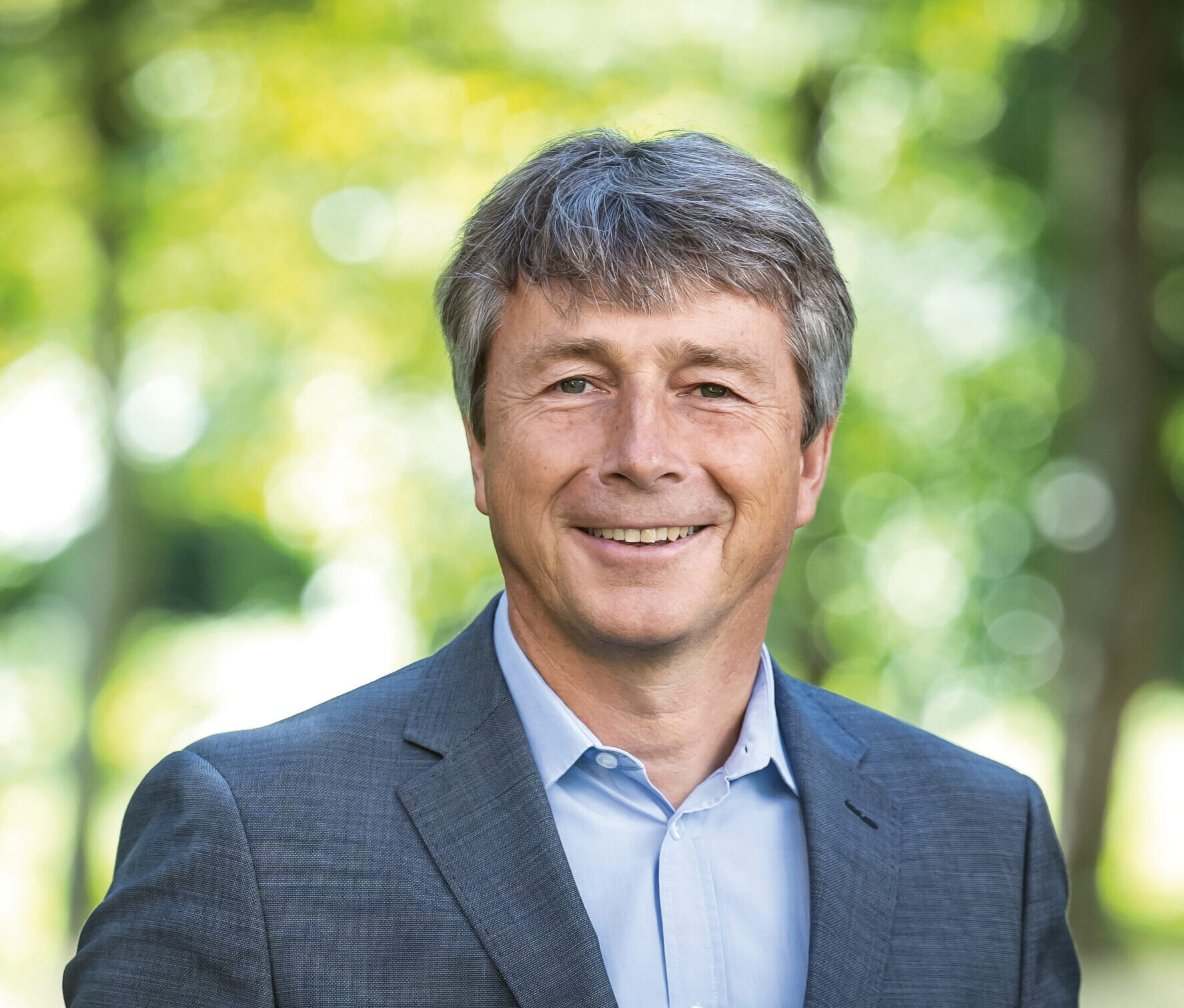 Lächelnder Mann in grauem Anzug und blauem Hemd, der im Freien vor einem unscharfen, grünen, grünen Hintergrund steht.