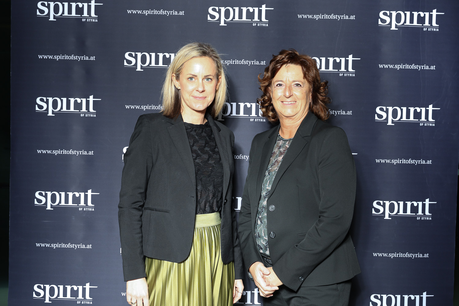 Zwei Frauen stehen lächelnd vor einem "spirit of styria" Step-and-Repeat-Hintergrund bei einer Veranstaltung.