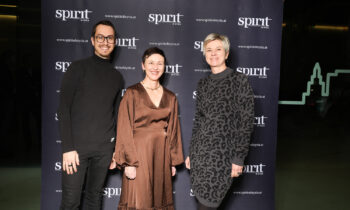 Drei Personen posieren und lächeln vor einem schwarzen Hintergrund mit "spirit of Styria"-Logos bei einer Veranstaltung.