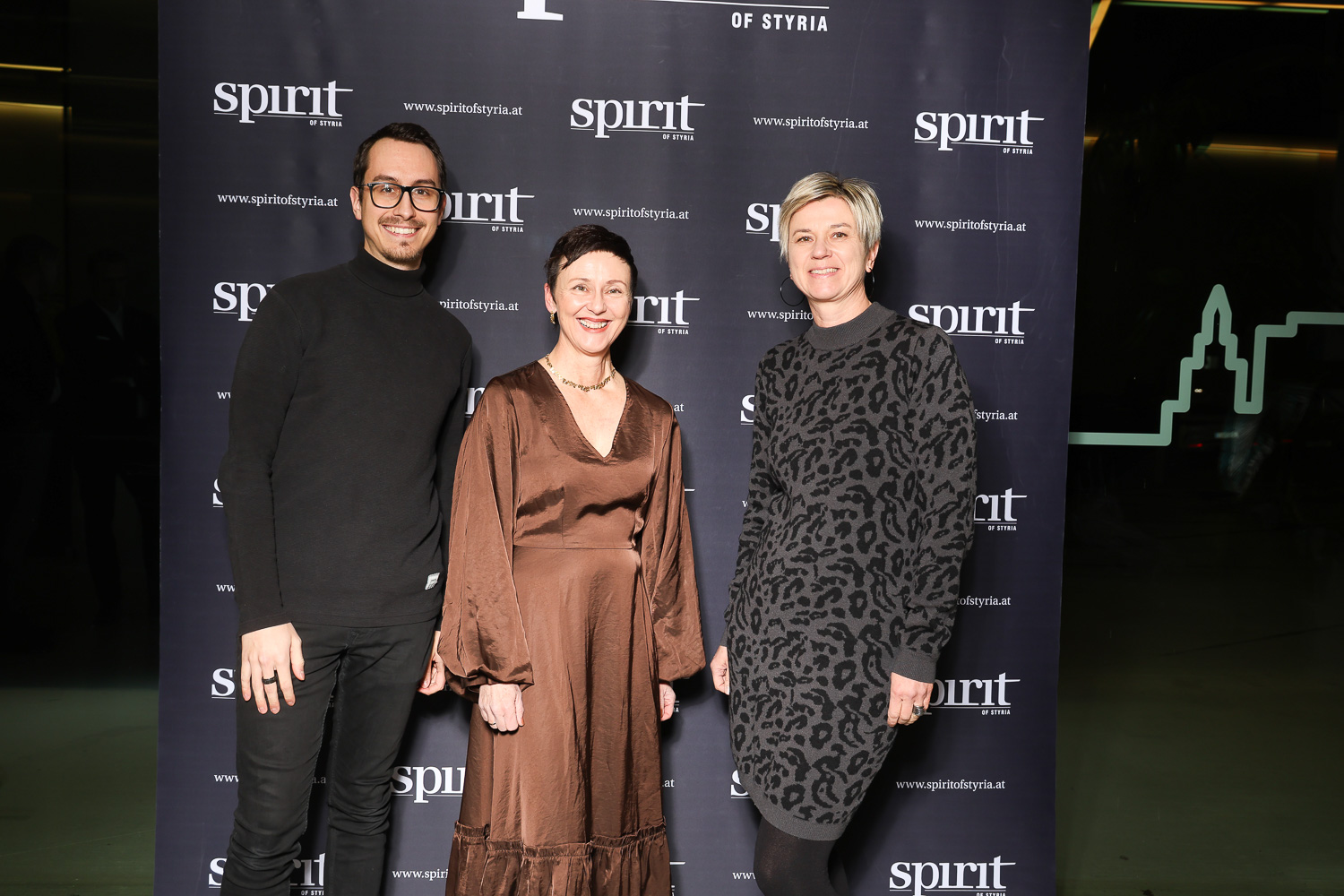 Drei Personen posieren und lächeln vor einem schwarzen Hintergrund mit "spirit of Styria"-Logos bei einer Veranstaltung.