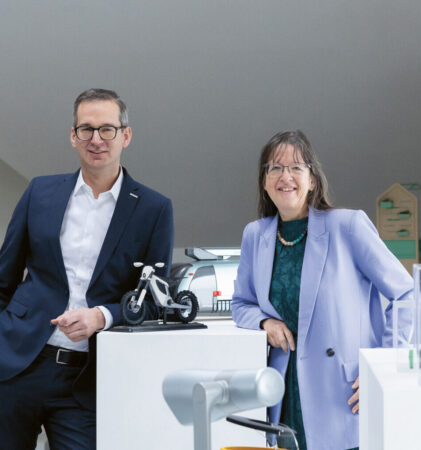 Zwei Personen in Business-Kleidung stehen in einem modernen, hellen Raum mit Holzbalken, umgeben von Technologie-Displays und innovativen Geräten.