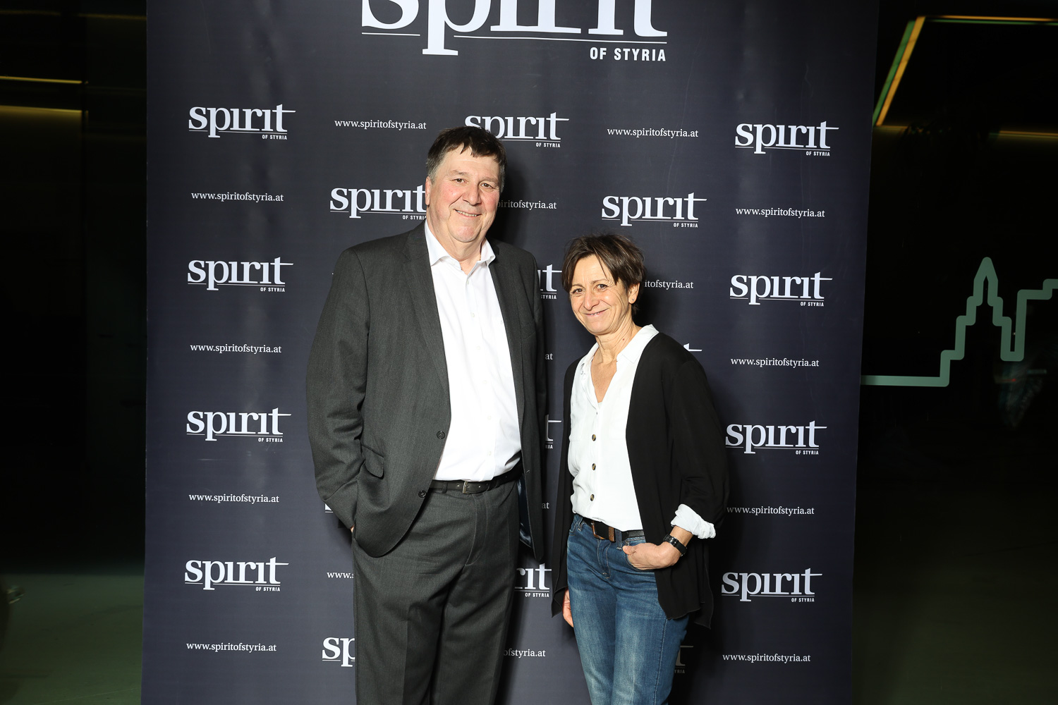 Zwei Personen stehen lächelnd vor einer Step-and-Repeat-Kulisse "Spirit of Styria" bei einer Veranstaltung.