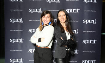 Zwei Frauen stehen Rücken an Rücken und lächeln in die Kamera, vor einem Hintergrund mit wiederholten "spirit of styria"-Logos.