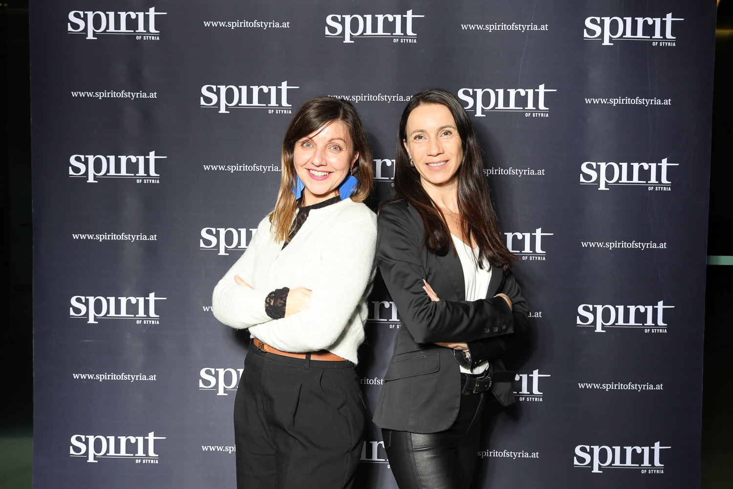 Zwei Frauen stehen Rücken an Rücken und lächeln in die Kamera, vor einem Hintergrund mit wiederholten "spirit of styria"-Logos.