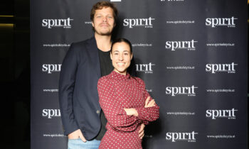 Ein Mann in einem dunklen Blazer und eine Frau in einem roten Tupfenkleid lächeln vor einem "Spirit of Styria"-Transparent im Step-and-Repeat-Verfahren.