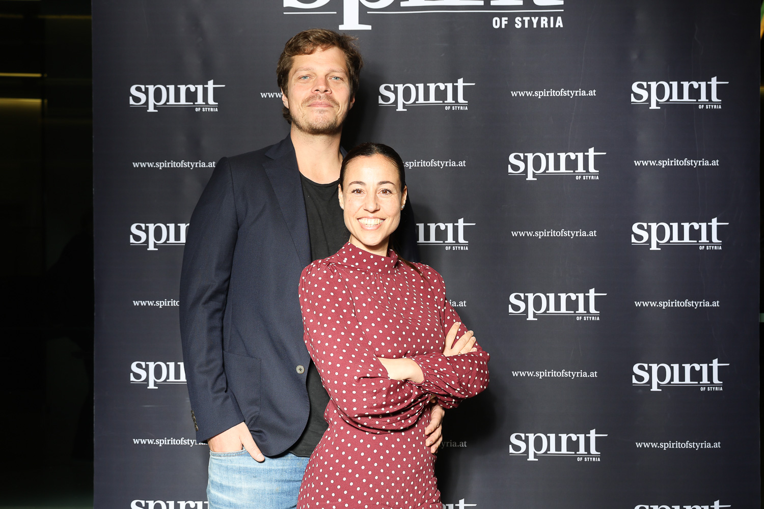 Ein Mann in einem dunklen Blazer und eine Frau in einem roten Tupfenkleid lächeln vor einem "Spirit of Styria"-Transparent im Step-and-Repeat-Verfahren.
