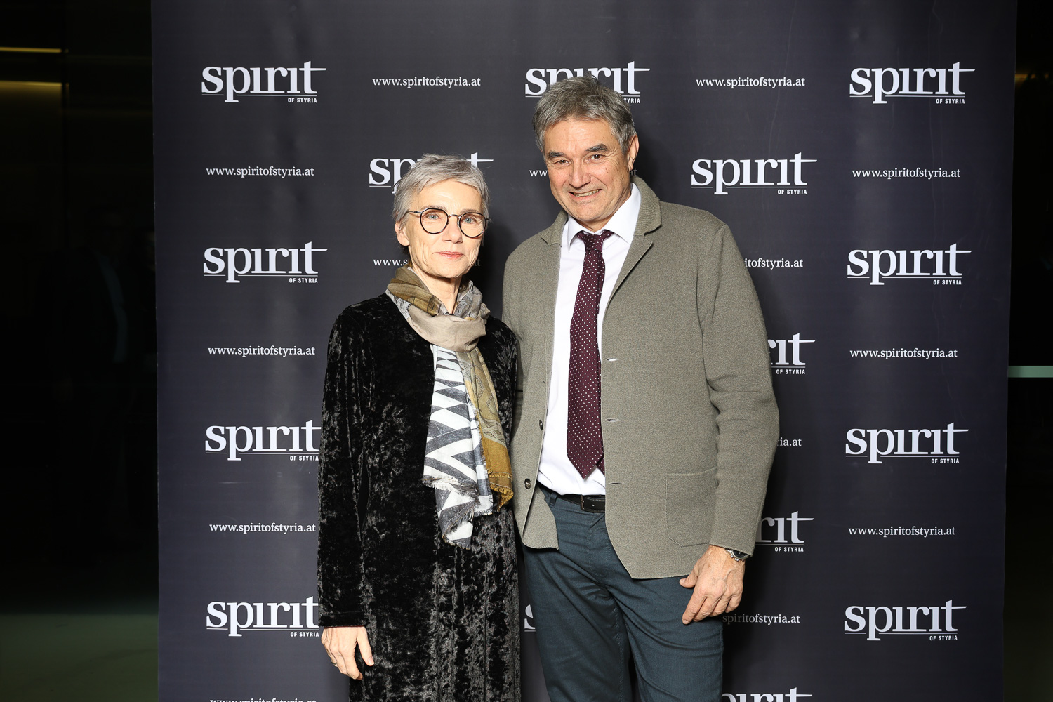 Eine Frau und ein Mann stehen bei einer Veranstaltung lächelnd vor einem schwarzen "Spirit of Styria"-Werbebanner.