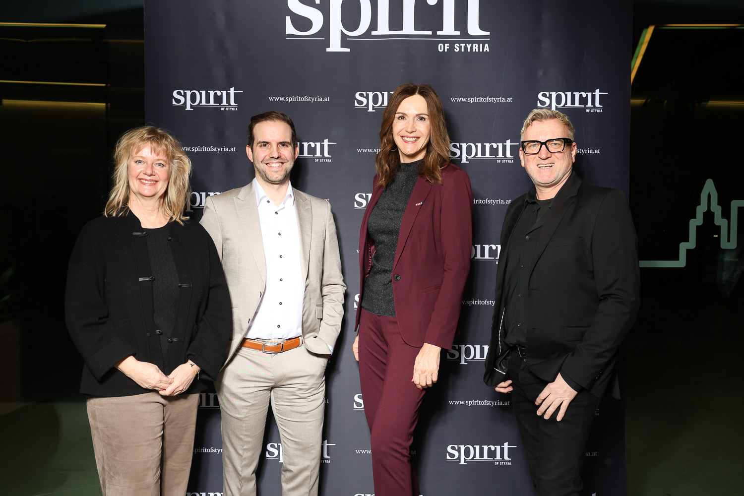 Vier Personen stehen lächelnd vor einem "spirit of styria"-Hintergrund bei einer Veranstaltung, gekleidet in Business- und Smart-Casual-Kleidung.