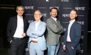Vier Männer in Business-Kleidung stehen bei einer Veranstaltung lächelnd vor einem schwarzen Step-and-Repeat-Banner "Spirit of Styria".