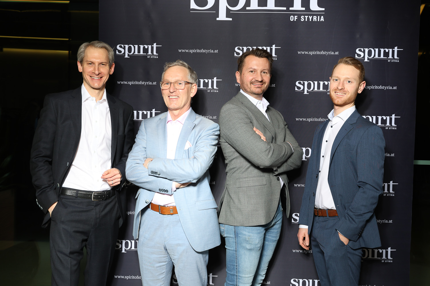 Vier Männer in Business-Kleidung stehen bei einer Veranstaltung lächelnd vor einem schwarzen Step-and-Repeat-Banner "Spirit of Styria".