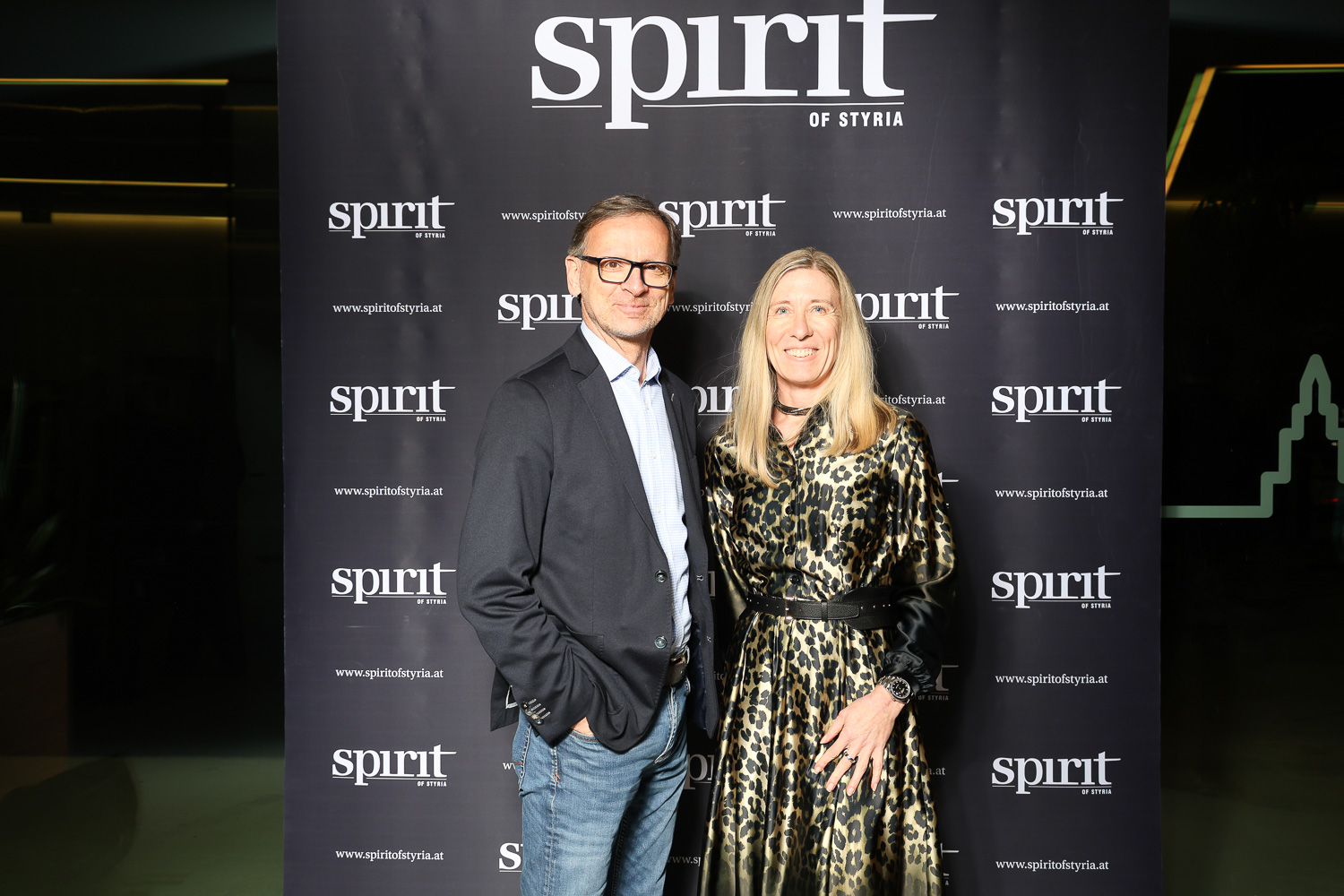 Ein Mann und eine Frau posieren gemeinsam lächelnd vor einer "Spirit of Styria"-Kulisse bei einer Veranstaltung.