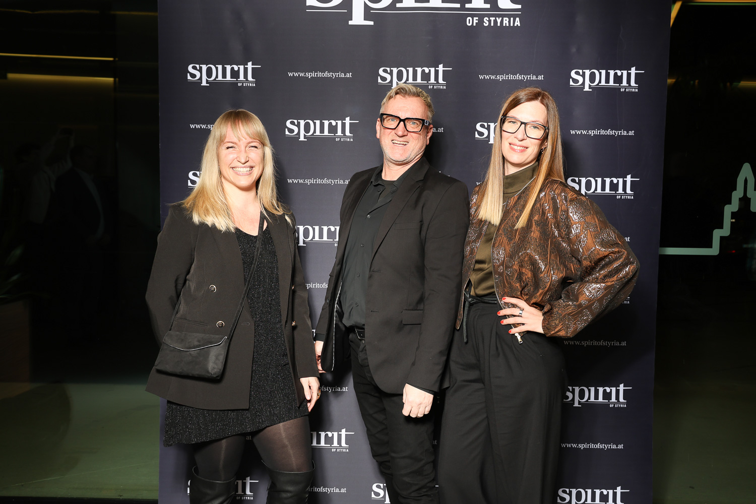 Drei Personen lächeln und posieren gemeinsam vor einem "Spirit of Styria" Step-and-Repeat-Banner bei einer Veranstaltung.