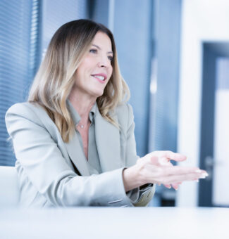 Eine Frau in einem hellen Blazer sitzt an einem Schreibtisch in einem modernen Büro, gestikuliert mit ihren Händen und lächelt während eines Gesprächs.