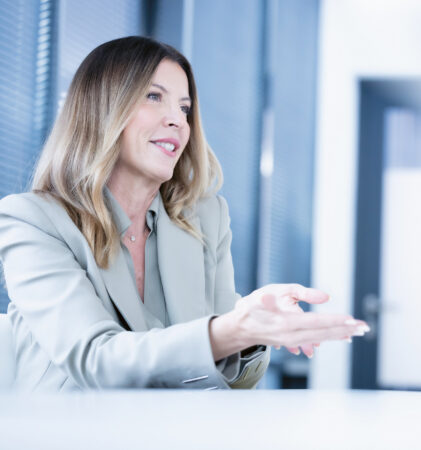Eine lächelnde Frau in einem hellen Anzug gestikuliert, während sie in einem modernen Büro mit großen Fenstern und natürlichem Licht spricht.