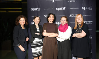 Fünf Frauen posieren und lächeln vor einer "Spirit of Styria"-Kulisse bei einer Veranstaltung, gekleidet in Business- und Semi-Formal-Kleidung.