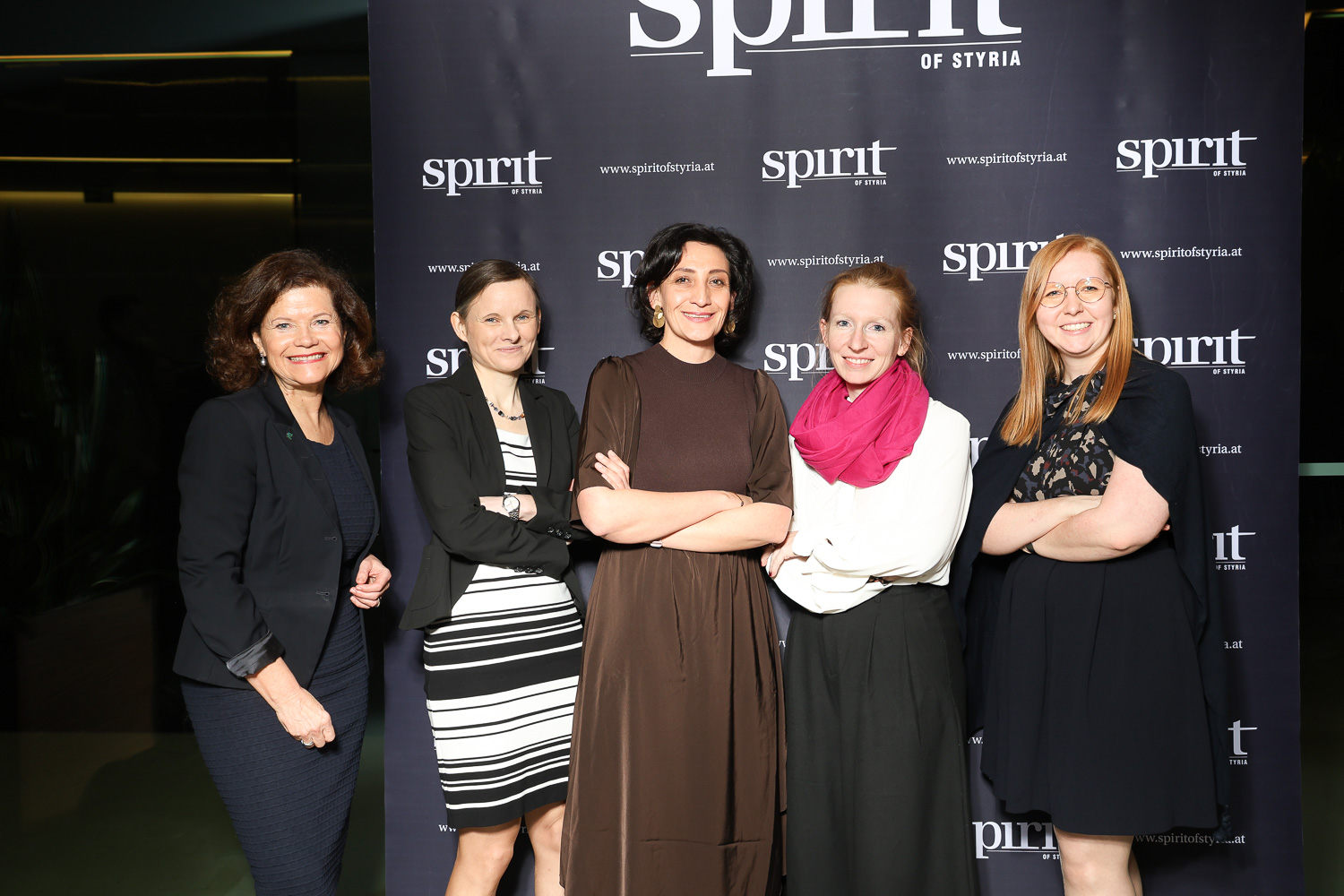 Fünf Frauen posieren und lächeln vor einer "Spirit of Styria"-Kulisse bei einer Veranstaltung, gekleidet in Business- und Semi-Formal-Kleidung.