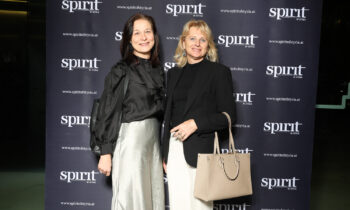 Zwei förmlich gekleidete Frauen stehen lächelnd vor einem schwarzen Step-and-Repeat-Hintergrund mit "spirit of styria"-Logos.