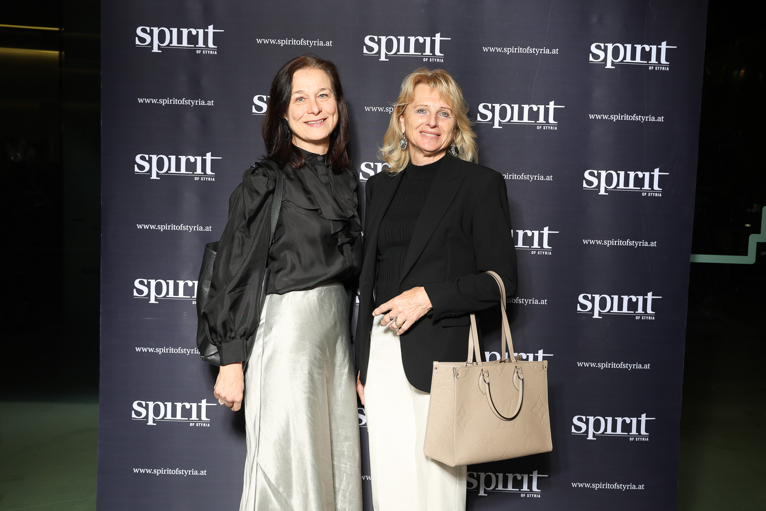 Zwei förmlich gekleidete Frauen stehen lächelnd vor einem schwarzen Step-and-Repeat-Hintergrund mit "spirit of styria"-Logos.