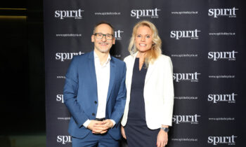 Zwei formal gekleidete Personen stehen lächelnd vor einem schwarzen Hintergrund mit wiederholten "spirit of Styria"-Logos.