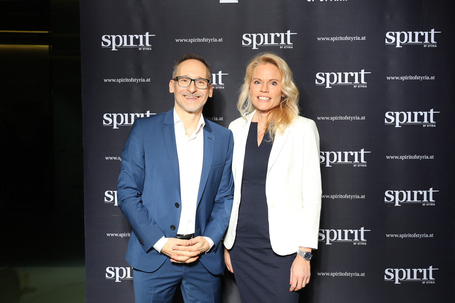 Zwei formal gekleidete Personen stehen lächelnd vor einem schwarzen Hintergrund mit wiederholten "spirit of Styria"-Logos.