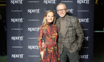 Eine Frau in einem gemusterten Kleid und ein Mann in einem karierten Anzug posieren und lächeln gemeinsam vor einem Step-and-Repeat-Banner mit "Spirit of Syria"-Logos.