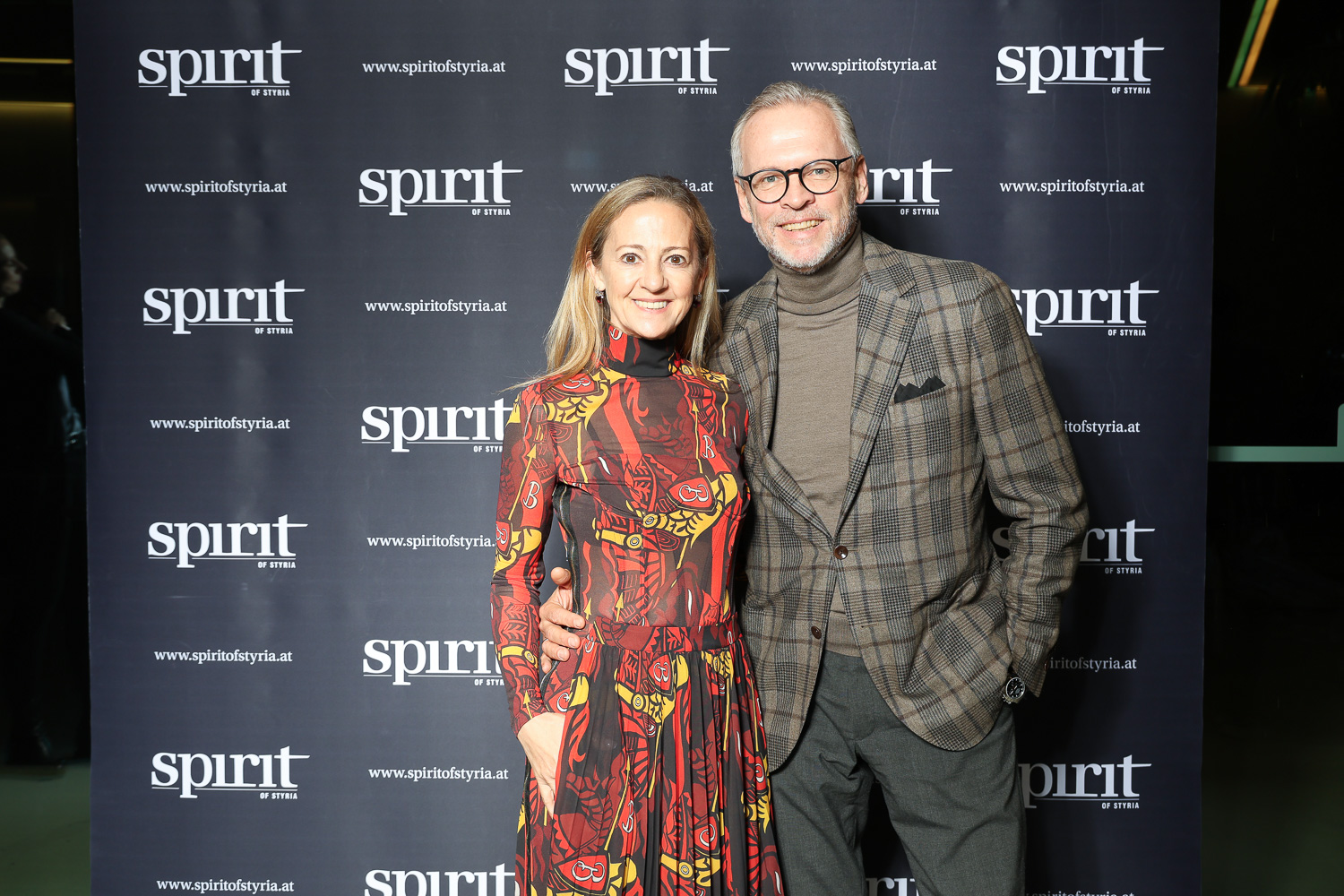 Eine Frau in einem gemusterten Kleid und ein Mann in einem karierten Anzug posieren und lächeln gemeinsam vor einem Step-and-Repeat-Banner mit "Spirit of Syria"-Logos.