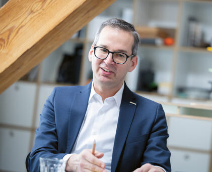 Ein Mann im Anzug und mit Brille sitzt an einem Schreibtisch, spricht und gestikuliert mit einem Bleistift in der Hand, während im Hintergrund Regale und Holzbalken zu sehen sind.