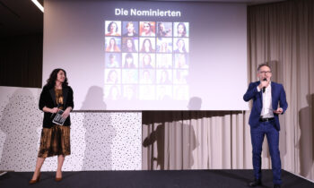 Eine Frau und ein Mann stehen auf der Bühne, hinter ihnen eine Leinwand mit Fotos der Nominierten und der Aufschrift "Die Nominierten".