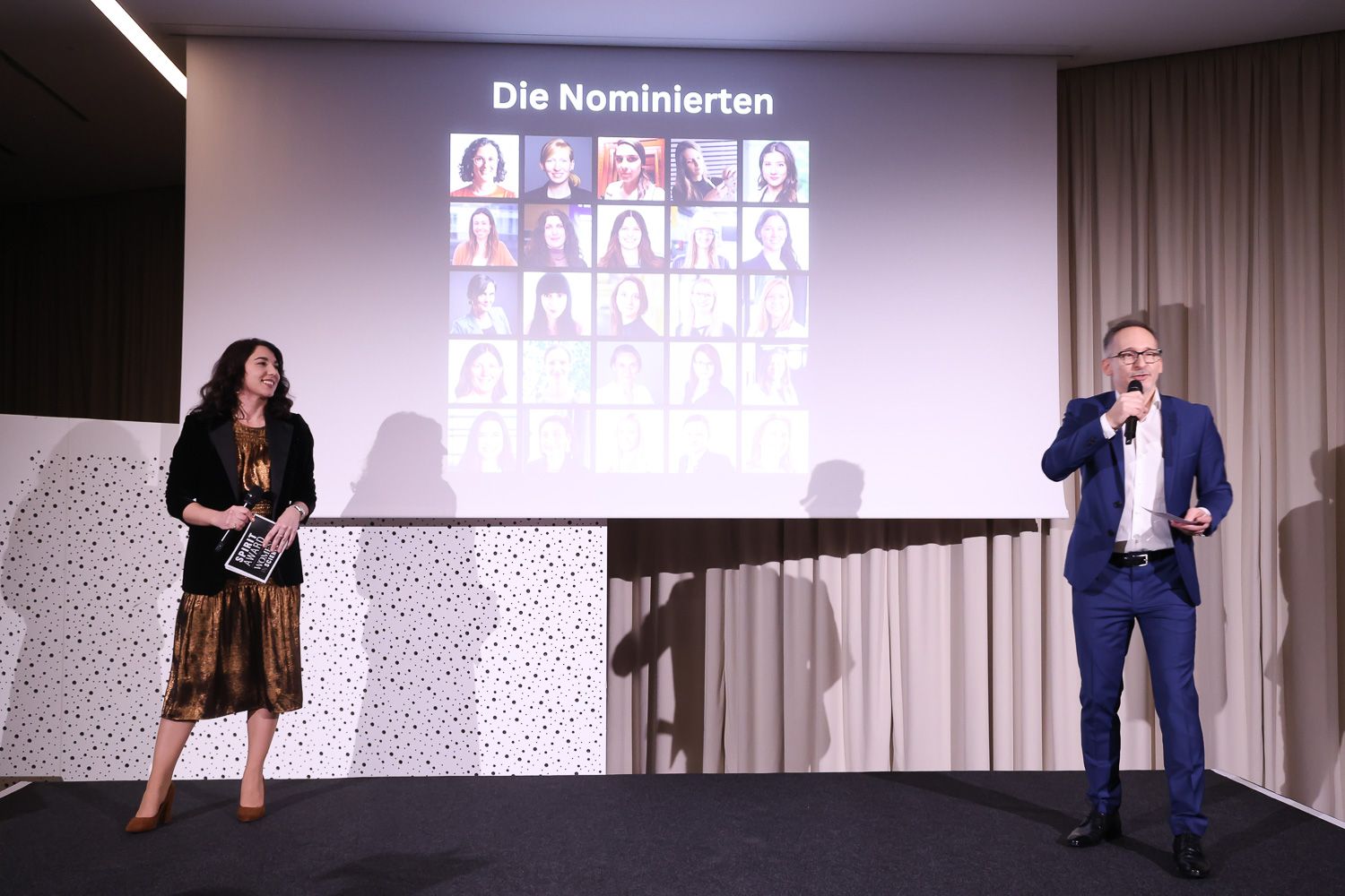 Eine Frau und ein Mann stehen auf der Bühne, hinter ihnen eine Leinwand mit Fotos der Nominierten und der Aufschrift "Die Nominierten".