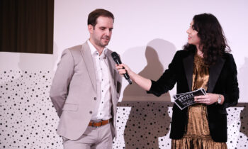 Eine Frau in einem goldenen Kleid und einem schwarzen Blazer hält einem Mann in einem hellen Anzug während einer Veranstaltung ein Mikrofon vor einem gepunkteten Hintergrund hin.