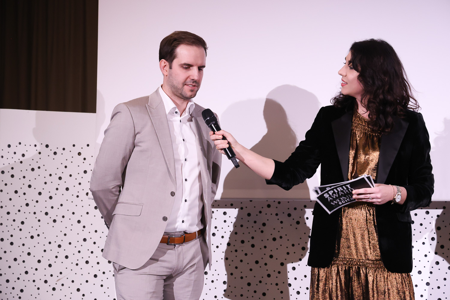 Eine Frau in einem goldenen Kleid und einem schwarzen Blazer hält einem Mann in einem hellen Anzug während einer Veranstaltung ein Mikrofon vor einem gepunkteten Hintergrund hin.