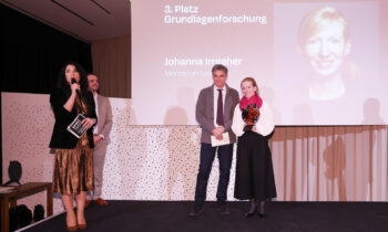 Vier Personen stehen auf der Bühne, eine Frau spricht in ein Mikrofon. Auf der Leinwand hinter ihnen erscheint "3. Platz Grundlagenforschung Johanna Irrgeher".