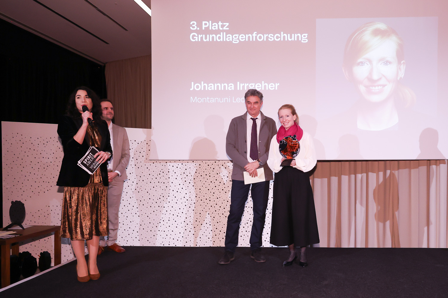 Vier Personen stehen auf der Bühne, eine Frau spricht in ein Mikrofon. Auf der Leinwand hinter ihnen erscheint "3. Platz Grundlagenforschung Johanna Irrgeher".