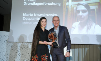 Zwei Personen stehen lächelnd auf der Bühne und halten einen Preis in der Hand. Hinter ihnen werden ein Bild und ein Text projiziert, die zeigen, dass Marta Nowakowska-Desplantes den zweiten Platz belegt hat.