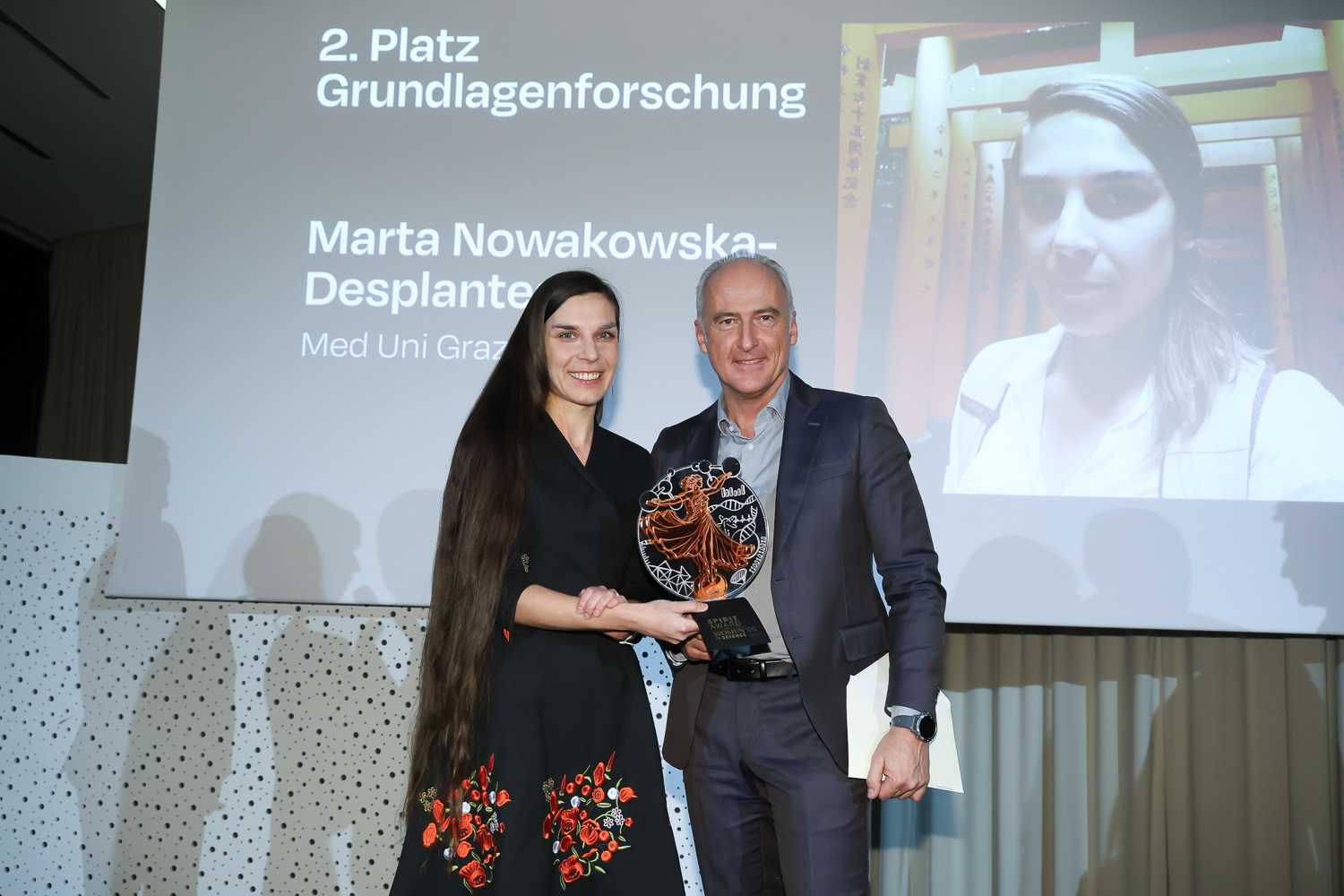 Zwei Personen stehen lächelnd auf der Bühne und halten einen Preis in der Hand. Hinter ihnen werden ein Bild und ein Text projiziert, die zeigen, dass Marta Nowakowska-Desplantes den zweiten Platz belegt hat.