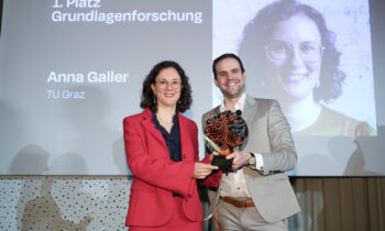 Zwei Personen lächeln und halten einen Preis in der Hand. Hinter ihnen zeigt ein Bildschirm "1. Platz Grundlagenforschung", "Anna Galler" und "TU Graz" mit einem großen Porträt.