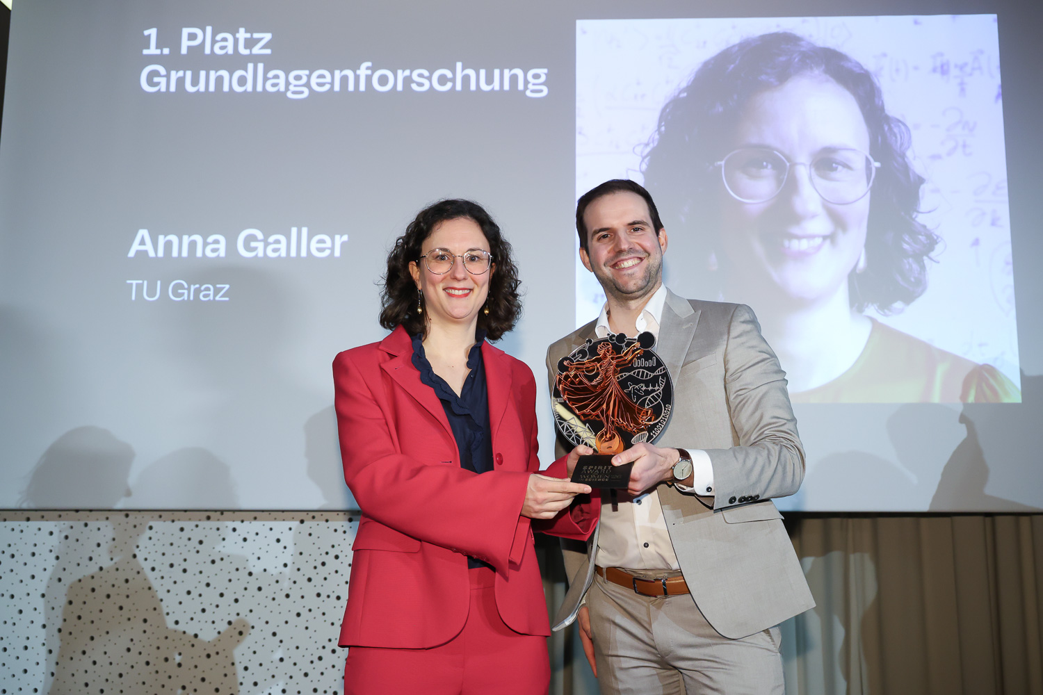Zwei Personen lächeln und halten einen Preis in der Hand. Hinter ihnen zeigt ein Bildschirm "1. Platz Grundlagenforschung", "Anna Galler" und "TU Graz" mit einem großen Porträt.