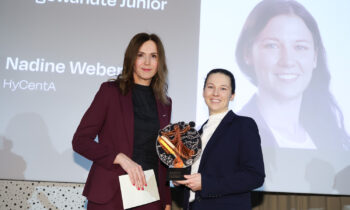 Zwei Frauen in formeller Kleidung posieren mit einer Auszeichnung; eine hält eine Trophäe. Eine Projektionsfläche im Hintergrund zeigt den Namen und das Porträt einer der Frauen.
