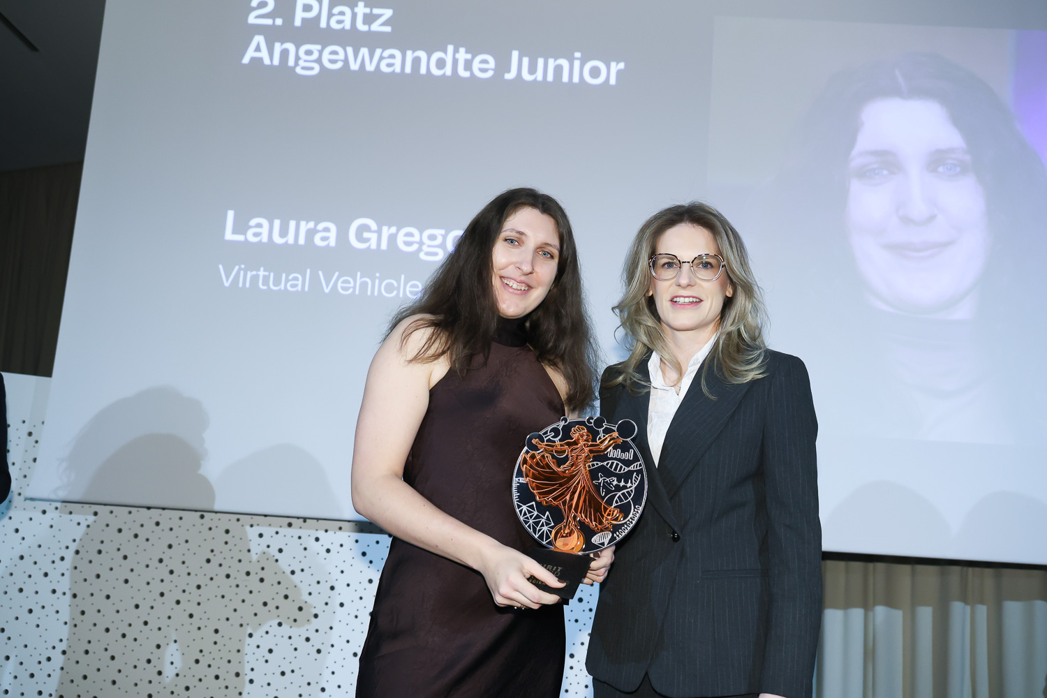 Zwei Frauen stehen lächelnd bei einer Preisverleihung. Eine von ihnen hält eine dekorative Trophäe; auf dem Bildschirm hinter ihnen steht "2. Platz Angewandte Junior Laura Gregorich".