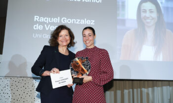 Zwei Frauen lächeln bei einer Preisverleihung; eine hält eine Plakette und eine Urkunde. Auf einem Bildschirm hinter ihnen werden "Raquel Gonzalez de Vesga" und ein Foto angezeigt.
