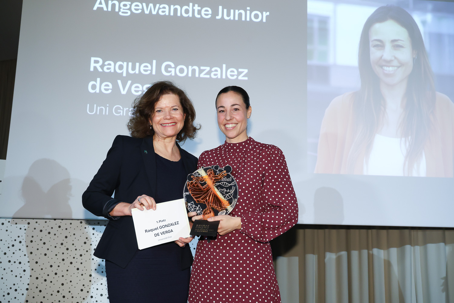 Zwei Frauen lächeln bei einer Preisverleihung; eine hält eine Plakette und eine Urkunde. Auf einem Bildschirm hinter ihnen werden "Raquel Gonzalez de Vesga" und ein Foto angezeigt.