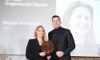 Zwei formell gekleidete Personen halten auf der Bühne einen Preis, hinter ihnen wird auf einer Leinwand "Nicole Wermuth" und "3. Platz Angewandte Senior" angezeigt.